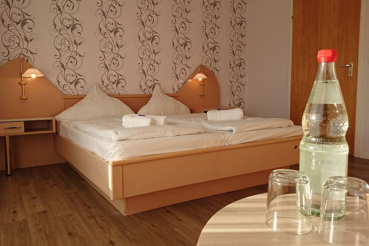 Un lit bien fait, avec du linge de lit blanc et deux oreillers, une table de chevet avec une lampe, et une bouteille d’eau posée sur le sol.