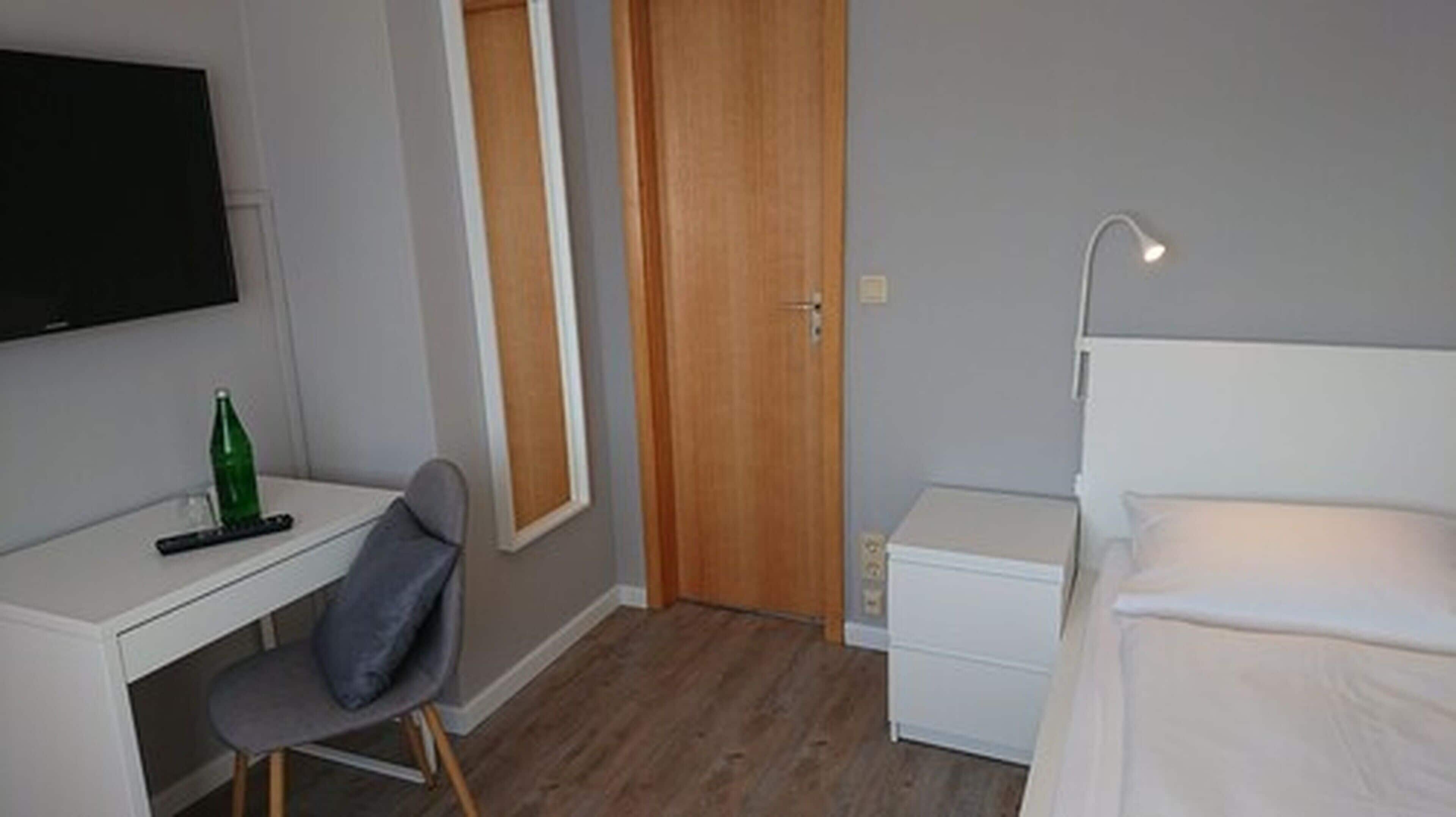 Quarto pequeno e bem organizado, com cama, escrivaninha, cadeira, televisão fixada na parede e uma porta.