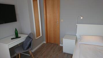 A small, neatly organized room with a bed, a desk, a chair, a TV mounted on the wall, and a door.
