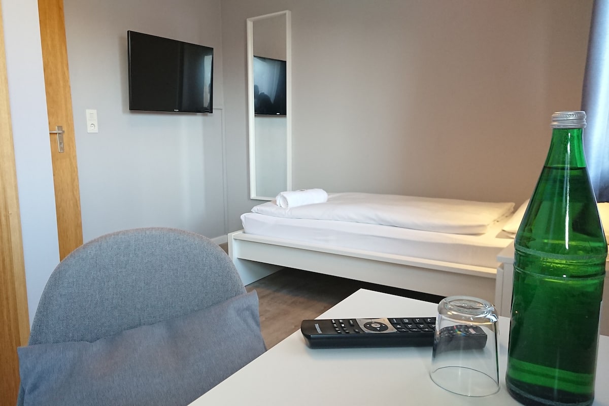 Une chambre d’hôtel avec un lit simple, un téléviseur fixé au mur, un bureau avec une télécommande, un verre et une bouteille verte.