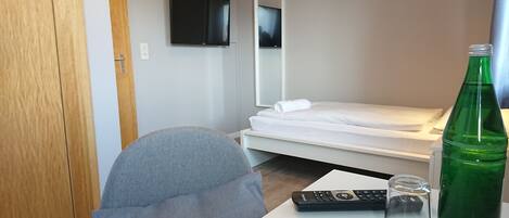 A hotel room with a single bed, a TV mounted on the wall, a desk with a remote control, a glass, and a green bottle.