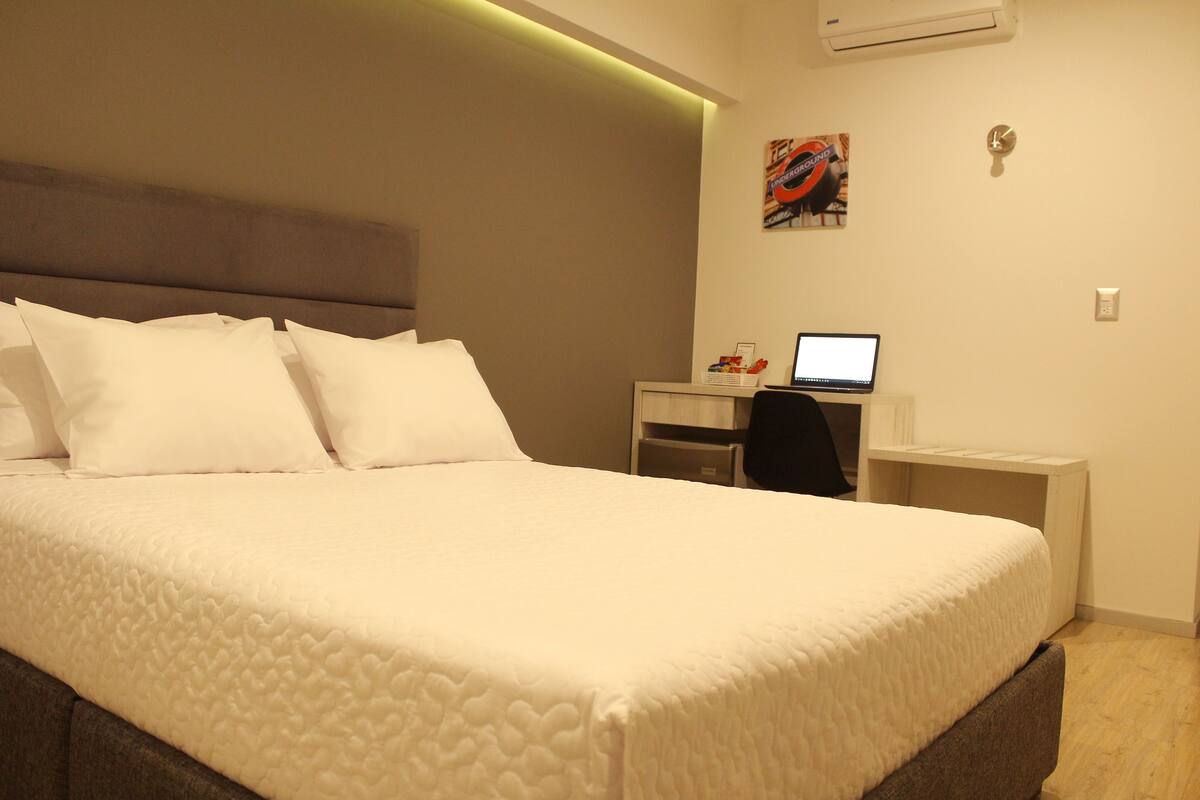 A neatly made bed with white linens, a small desk with a computer, and a wall-mounted air conditioner.