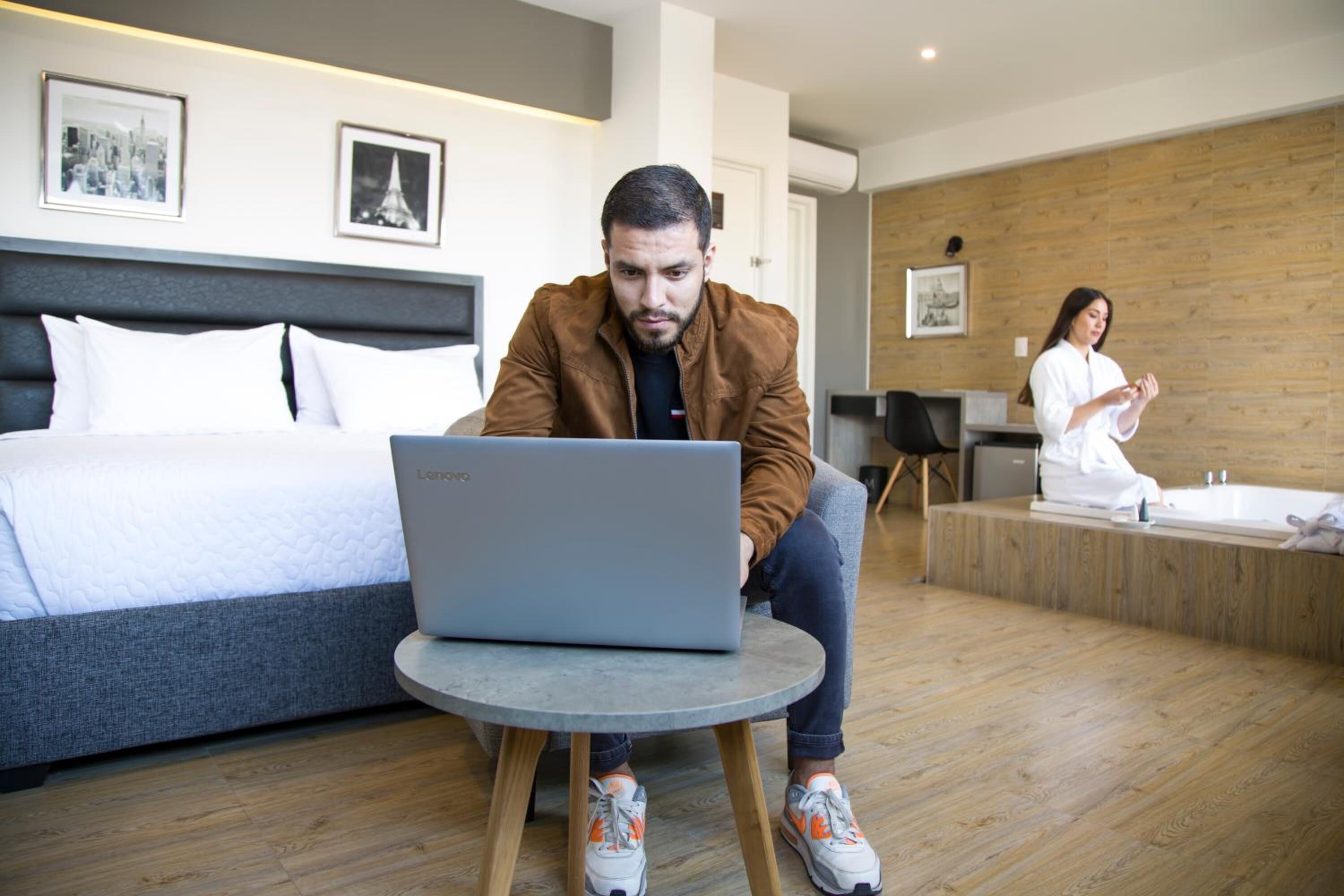 Une chambre d’hôtel moderne avec un lit, un ordinateur portable posé sur une petite table et une personne assise à un bureau.