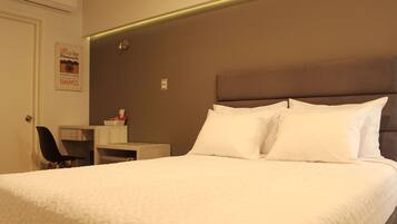 A neatly made bed with white bedding, a small desk with a chair, and a wall-mounted air conditioner.