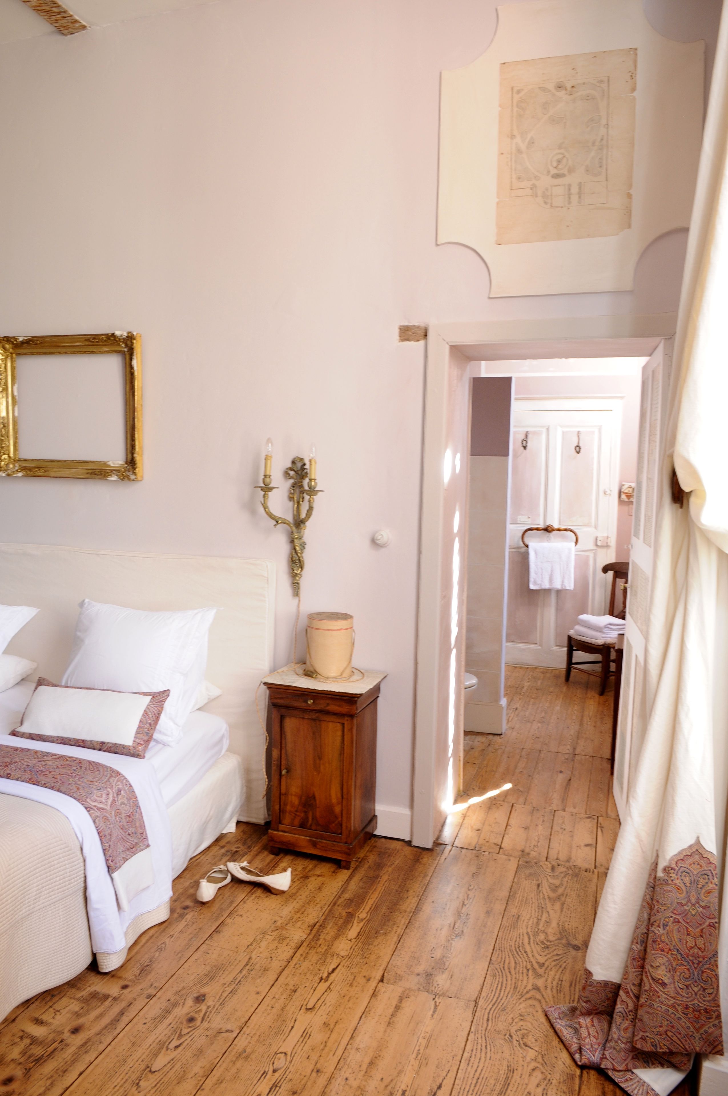 A bedroom with a wooden floor, a bed with white bedding, a small wooden nightstand, and a mirror with a gold frame.