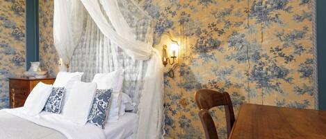 Un dormitorio con cama con dosel, papel tapiz floral, una mesita de noche de madera y una mesa de madera.