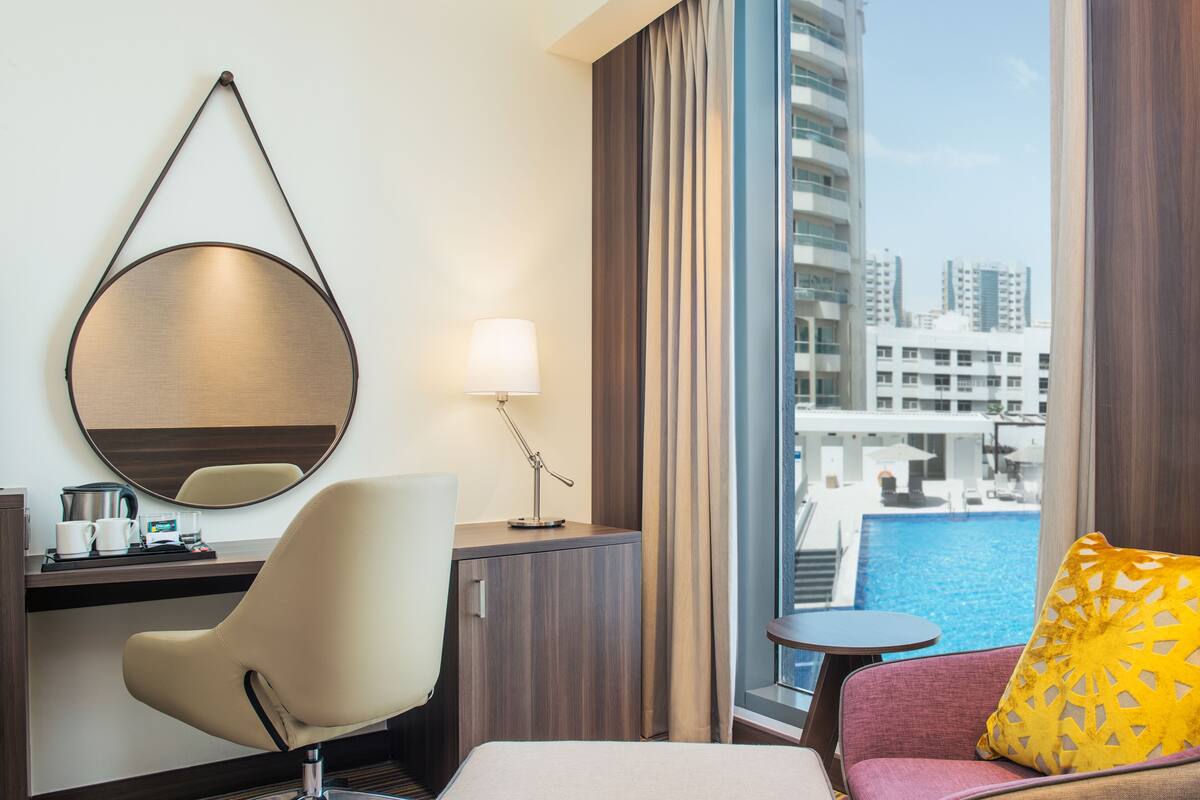 Une chambre d’hôtel avec un lit, un bureau doté d’un miroir, une chaise, une petite table, une lampe et une vue sur une piscine et des immeubles.