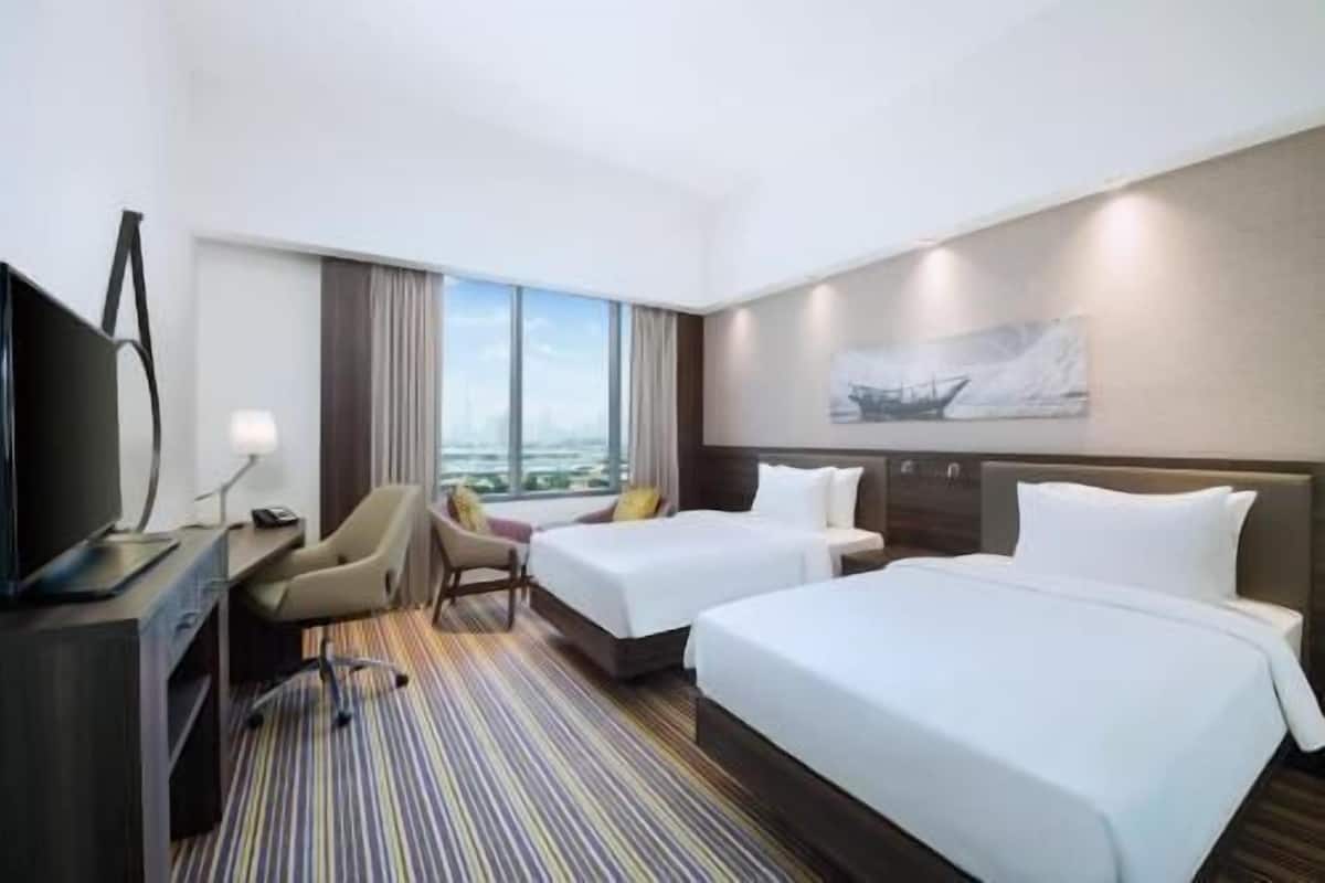 Une chambre d’hôtel avec deux lits, un bureau avec une télévision, une chaise et une grande fenêtre donnant sur la ville.