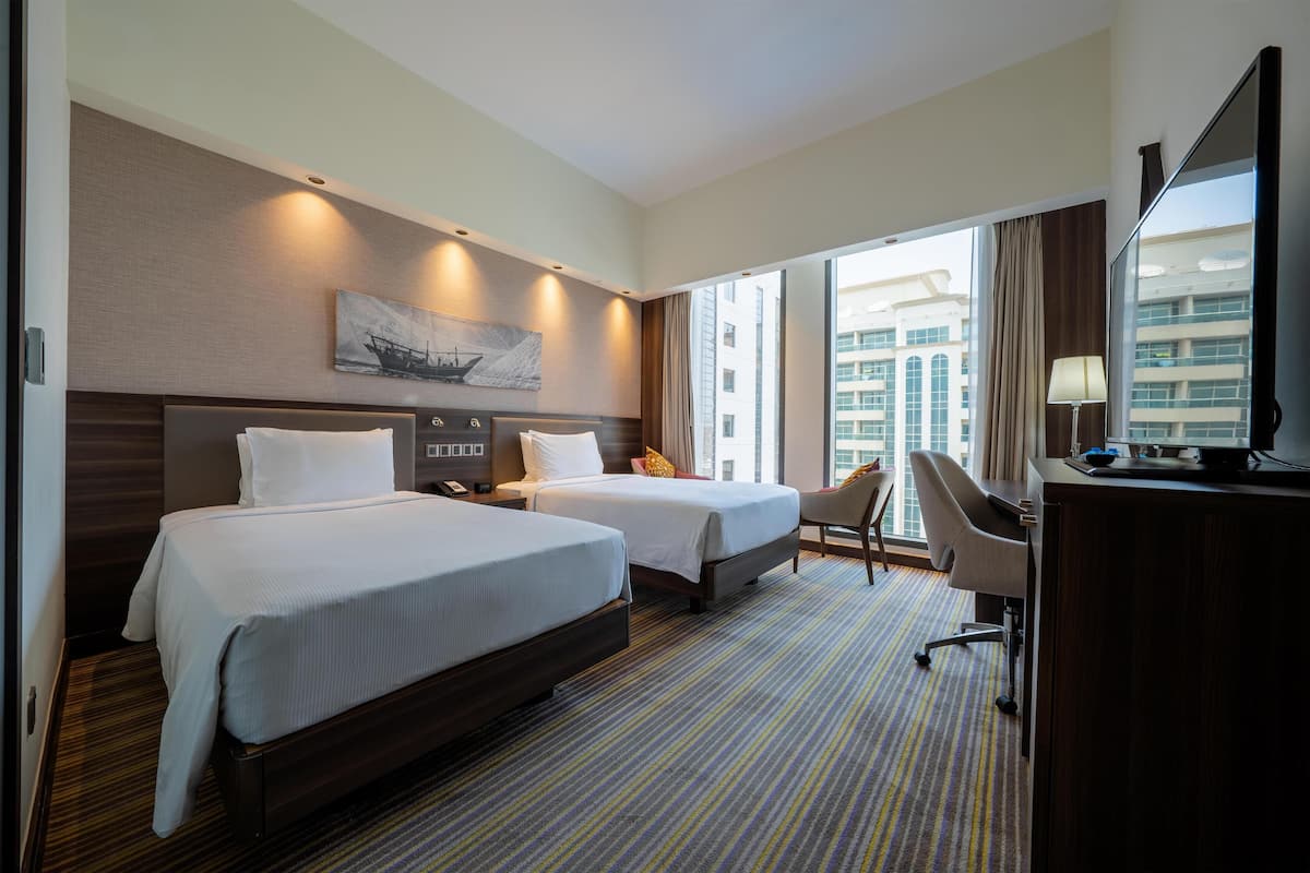 Une chambre d’hôtel avec deux lits, un bureau, une chaise, une télévision et une grande fenêtre donnant sur la ville.