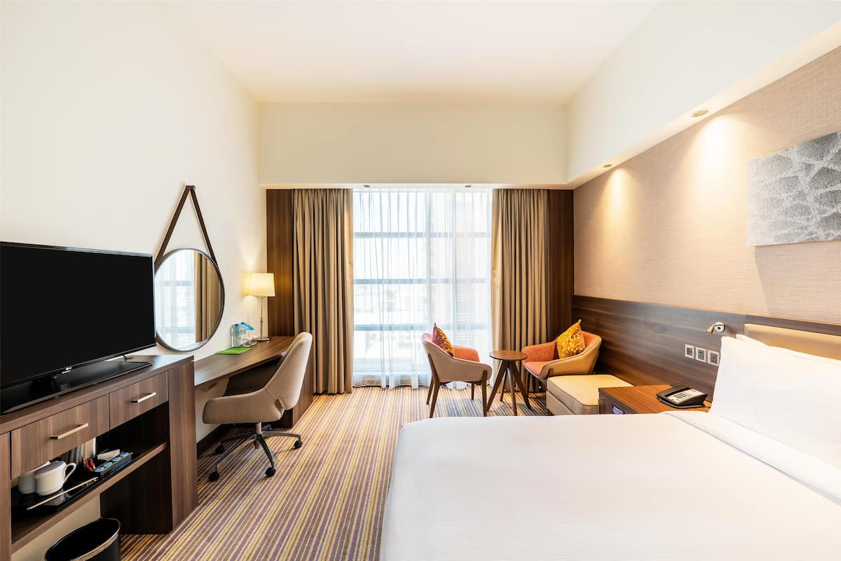 Une chambre d’hôtel avec un lit, un bureau avec une télévision, une chaise, une petite table et une fenêtre avec des rideaux.