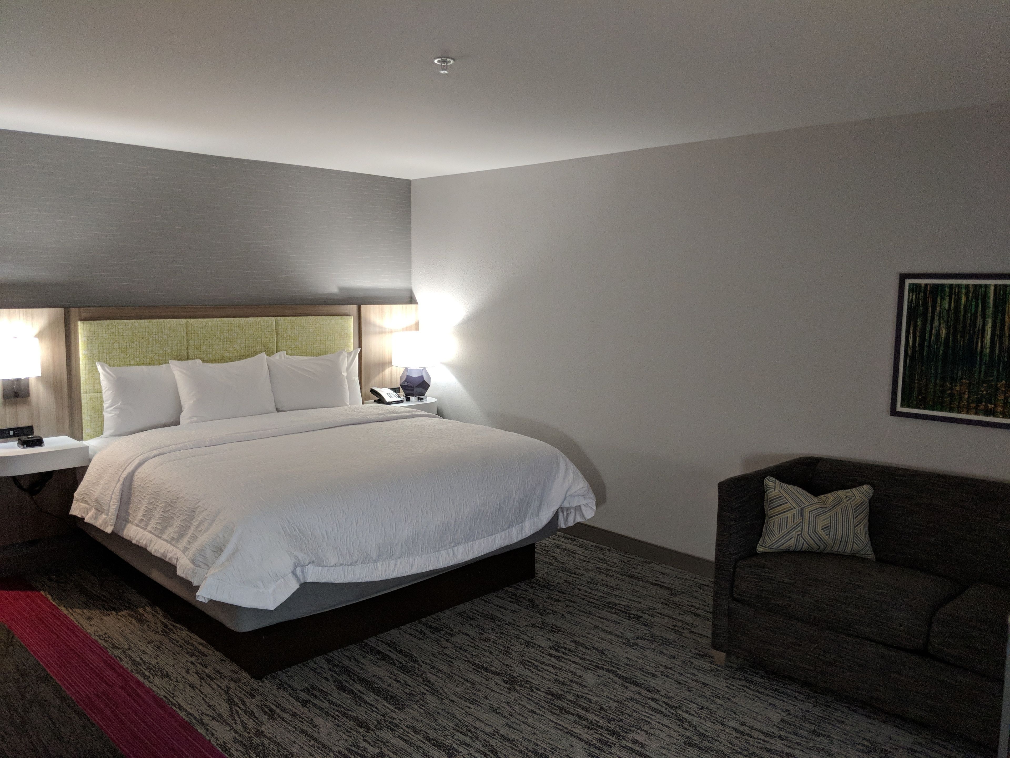 A hotel room with a large bed, a bedside table with a lamp, a small table with a phone, a dark sofa, and a framed picture on the wall.