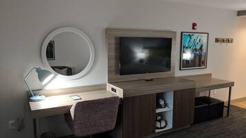 A hotel room with a wooden desk, a round mirror, a flat-screen TV, a chair, and a shelf with toiletries.