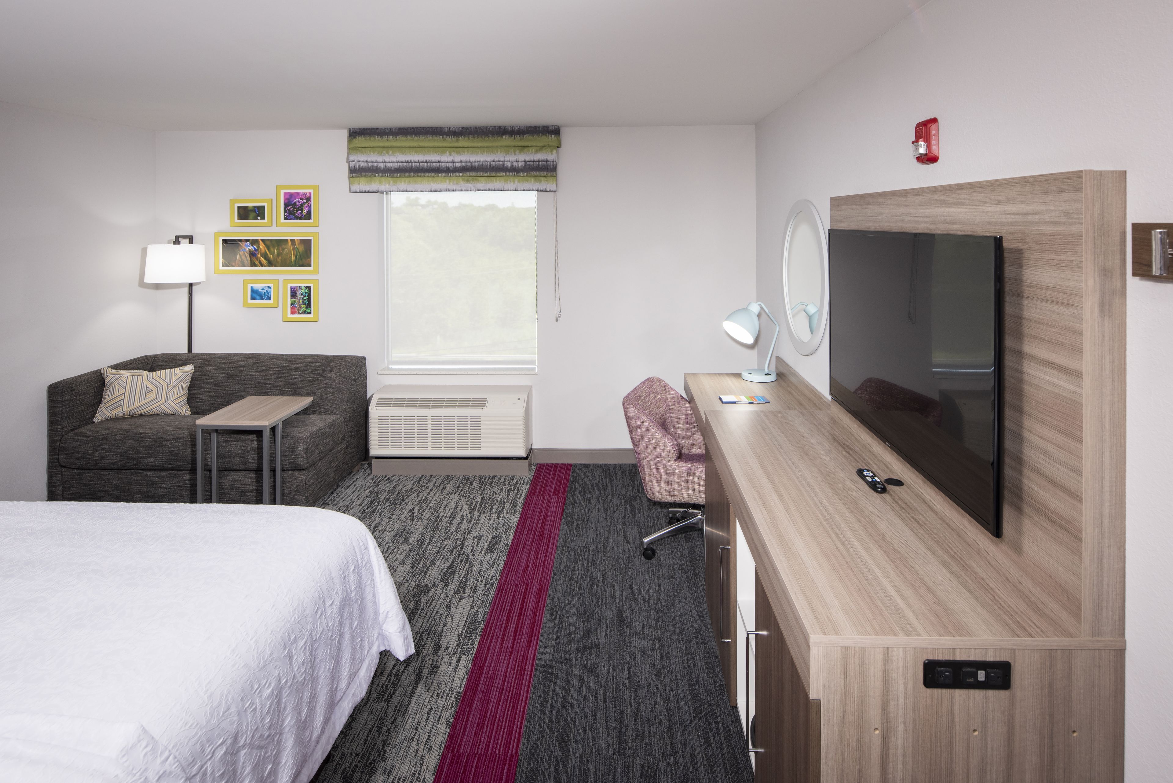 Quarto de hotel com cama, sofá, escrivaninha, televisão, abajur, espelho e janela com vista.