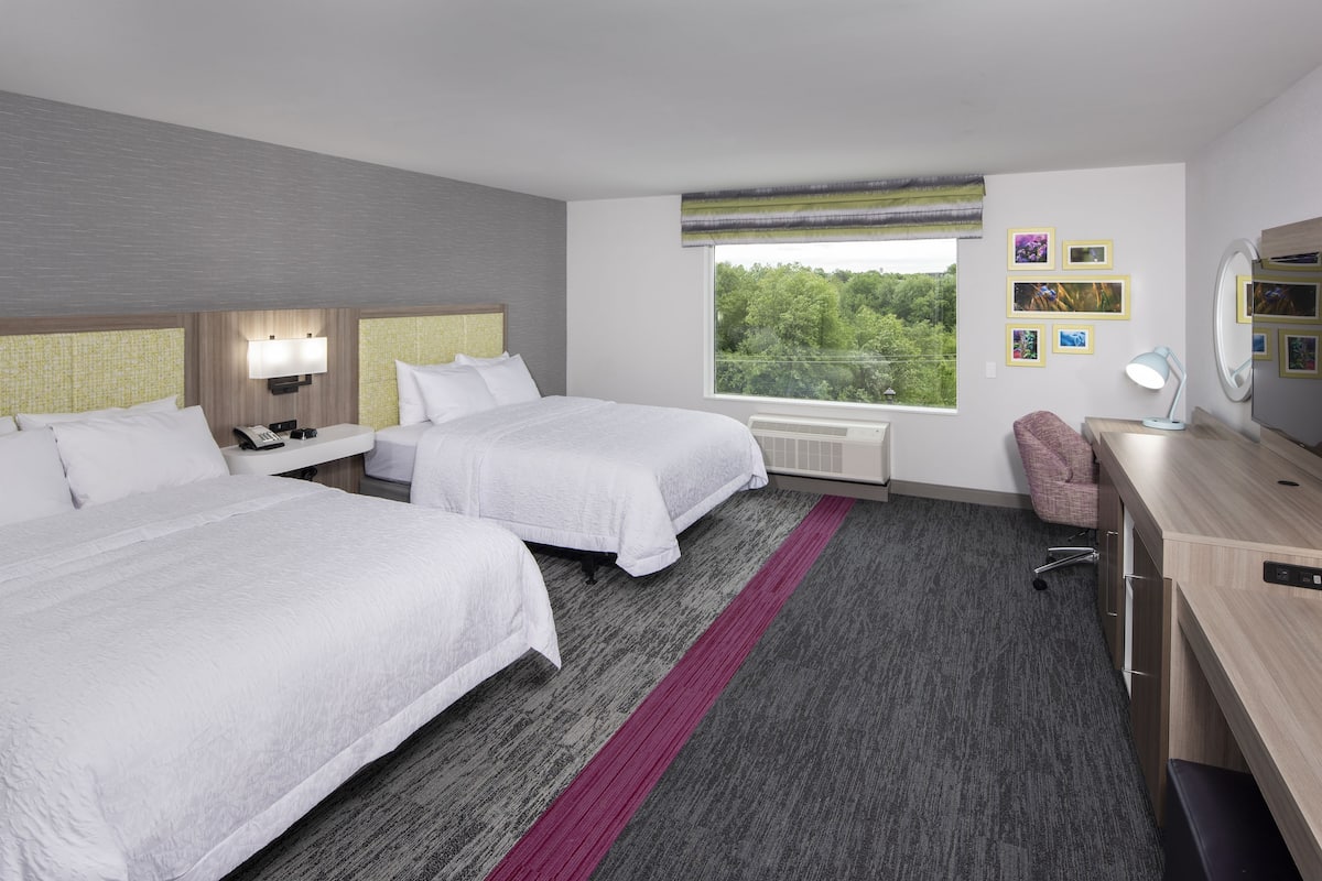 Habitación de hotel con dos camas, un escritorio, una silla, un ventanal con vistas y una pared decorada con cuadros.
