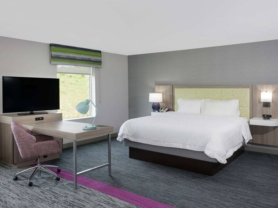 A hotel room with a bed, a desk with a TV, a chair, a window, and a nightstand with lamps.