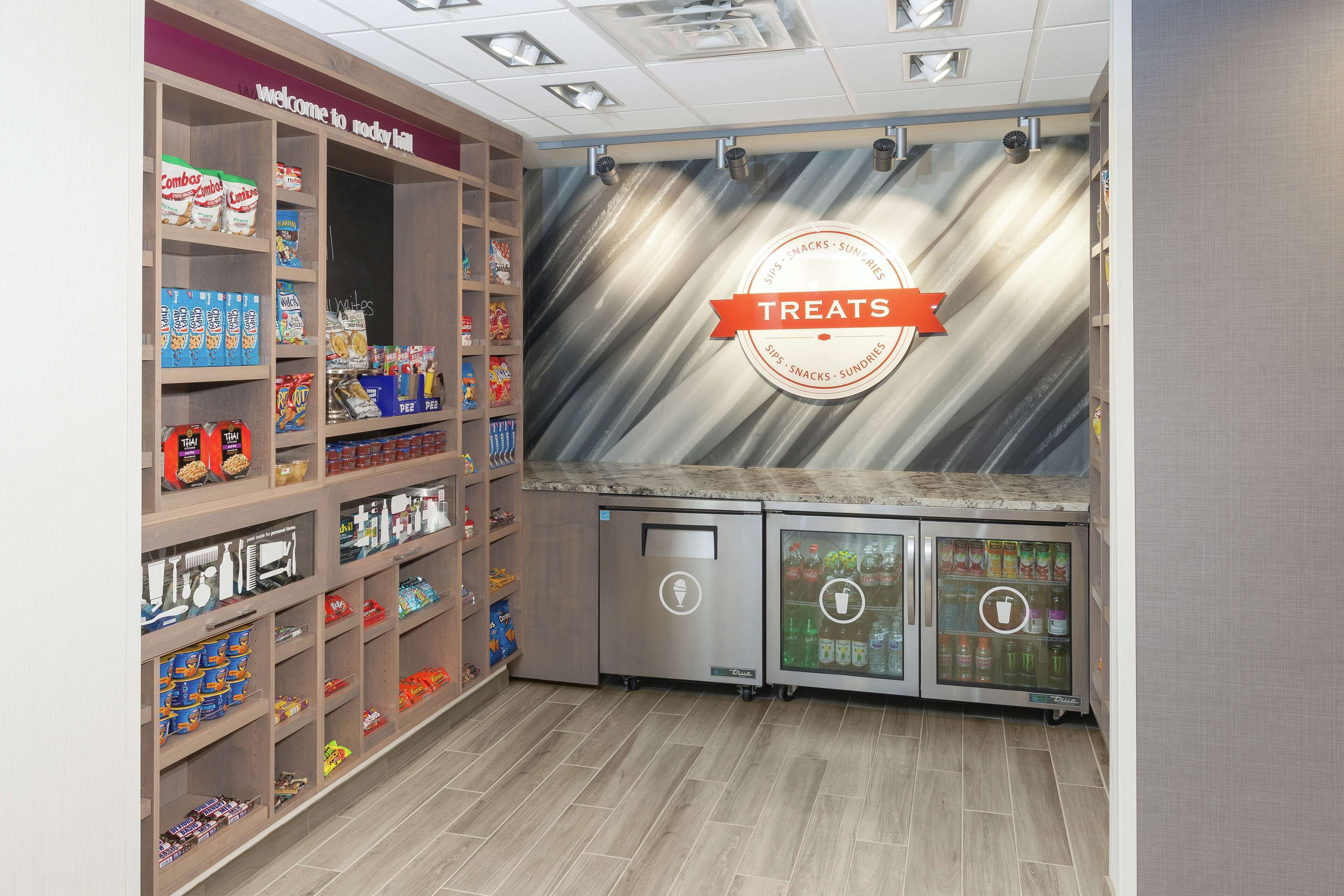 A snack bar with shelves stocked with various snacks and drinks, a refrigerated display case, and a 'Treats' sign on the wall.