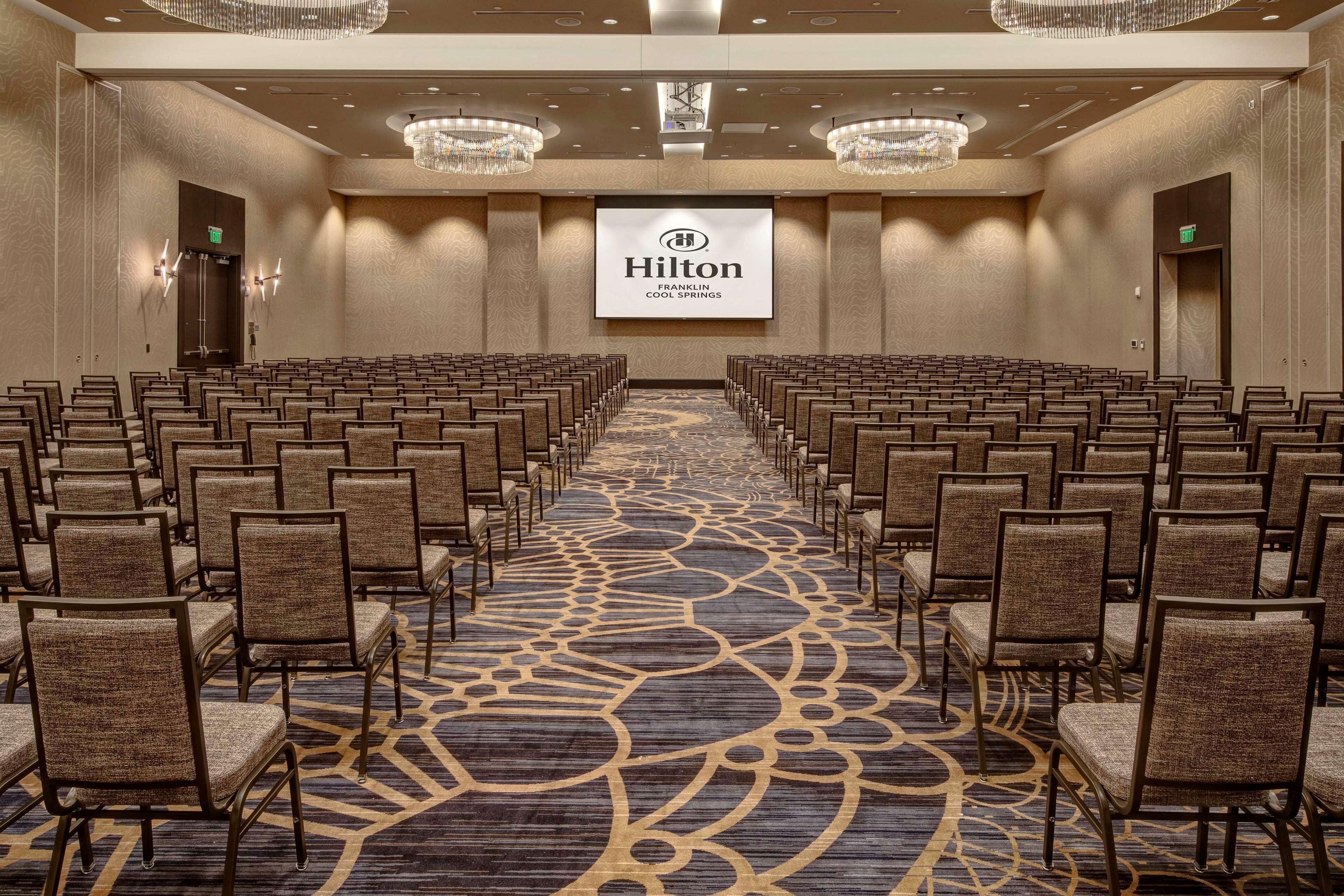 Ein Konferenzraum im Hilton Franklin Cool Springs mit Reihen von Stühlen, einem gemusterten Teppich, Kronleuchtern und einem großen Bildschirm, auf dem das Hilton-Logo zu sehen ist.