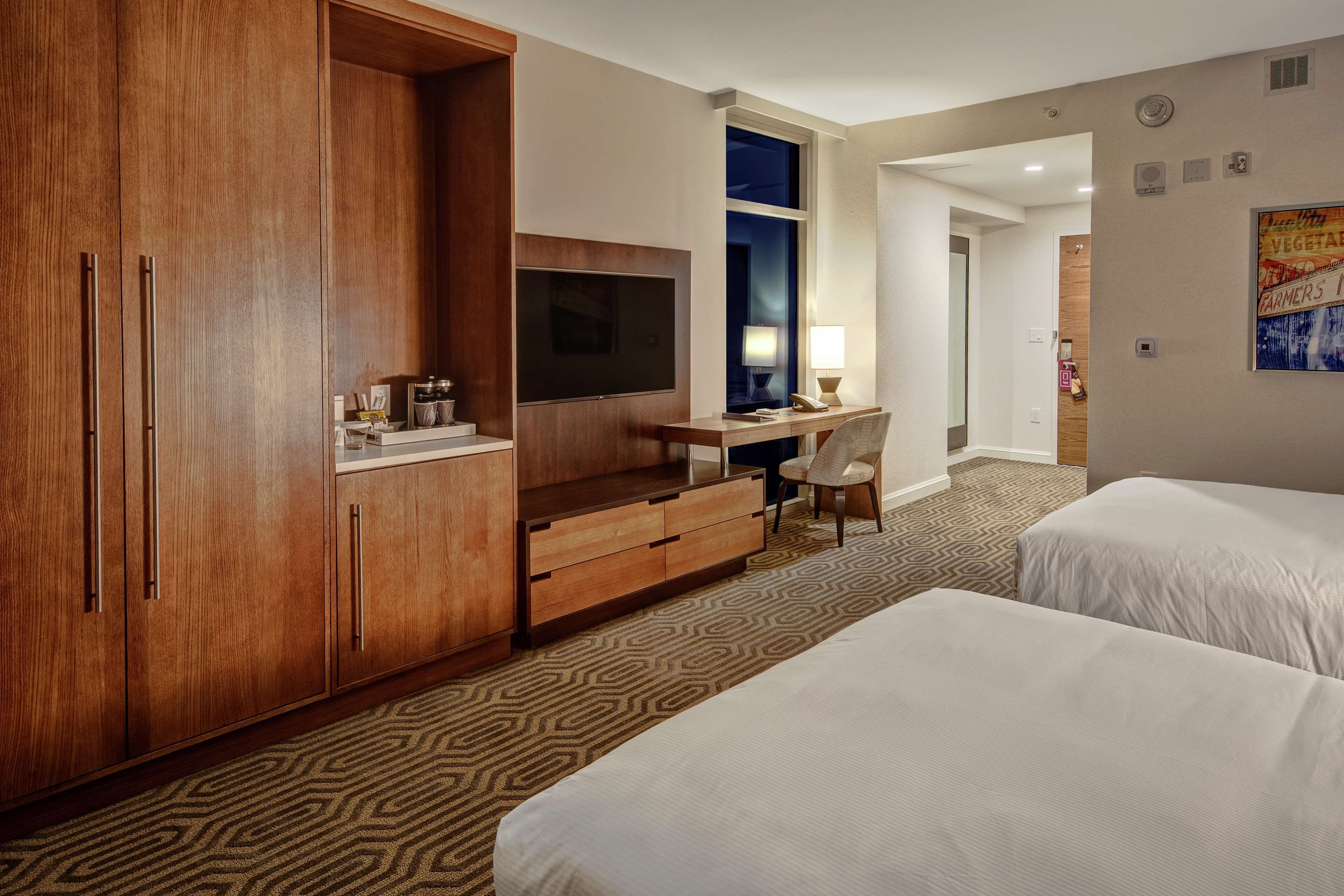 A hotel room with two beds, a wooden wardrobe, a desk with a chair, a television, and a patterned carpet.