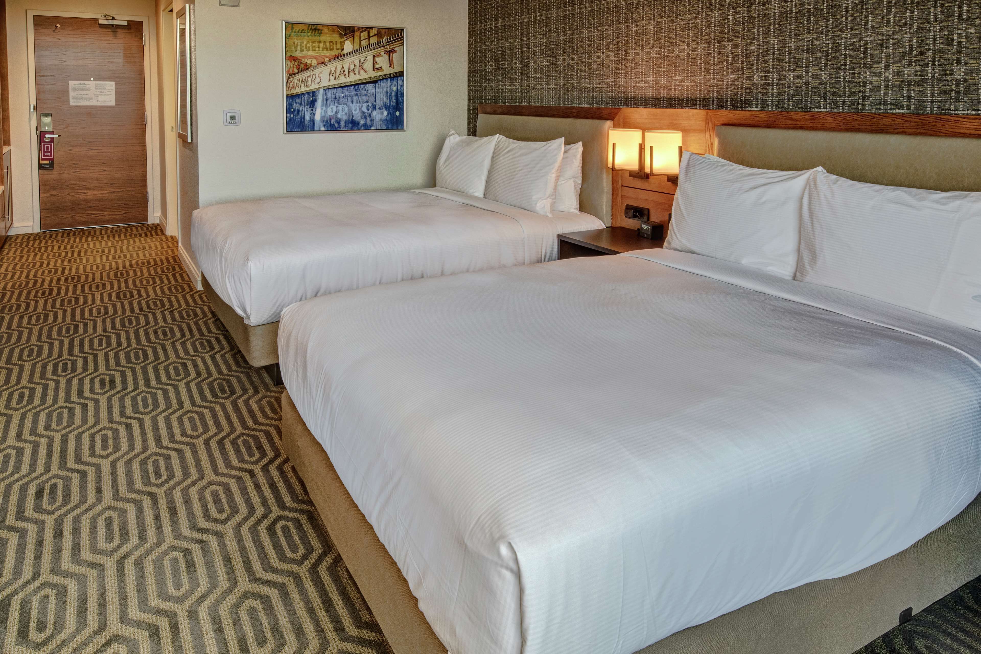 A hotel room with two beds, a patterned carpet, a wall clock, and a framed poster.