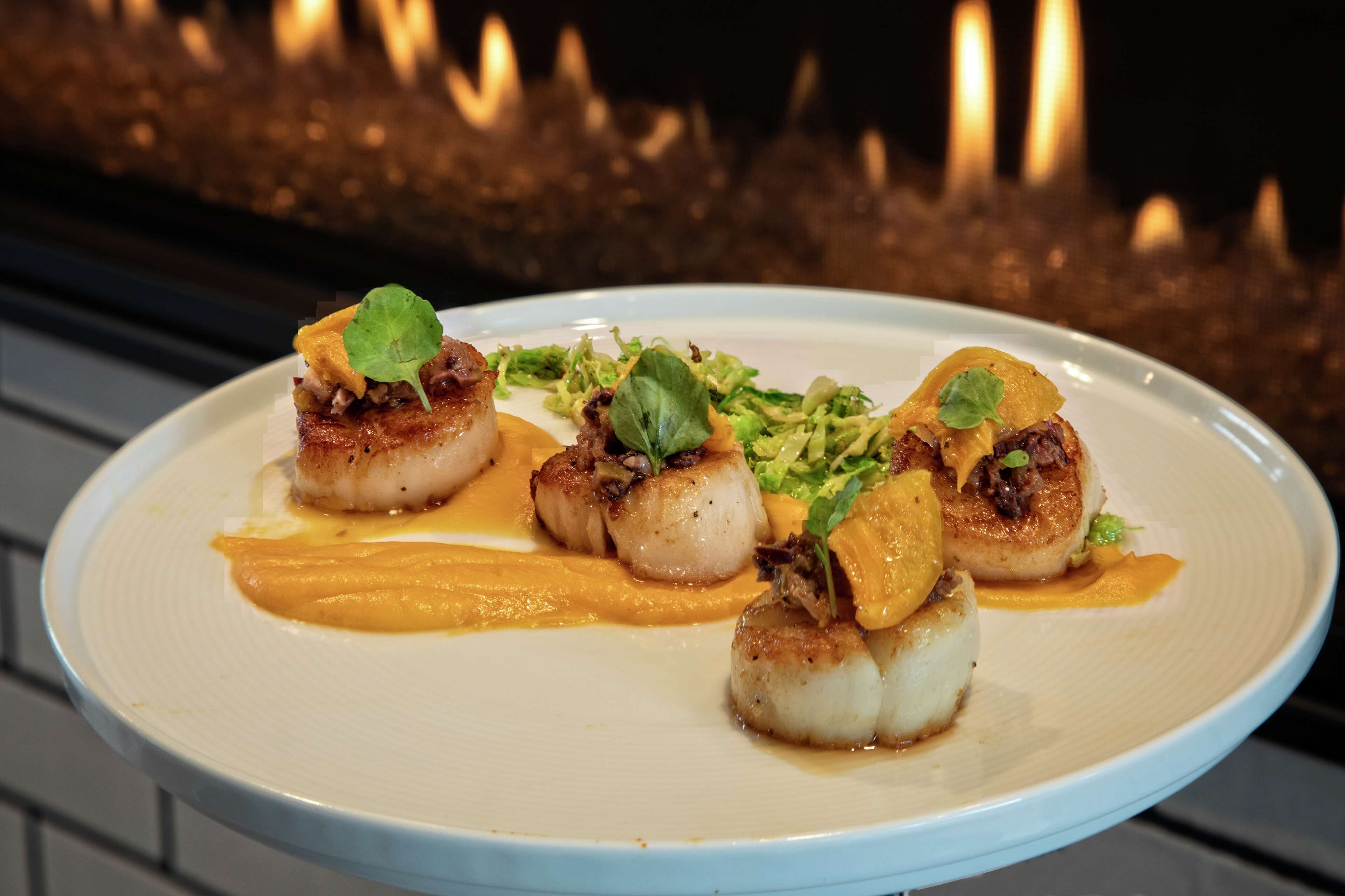 A plate of seared scallops with a creamy sauce and garnishes, set against a background of a lit fireplace.