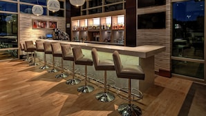 A bar area with multiple stools, a counter with bottles, and a large window. - Hilton Franklin Cool Springs (Franklin)