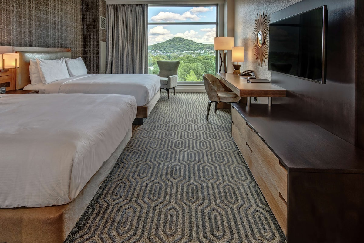 A hotel room with two beds, a desk, a TV, and a view of a mountain.