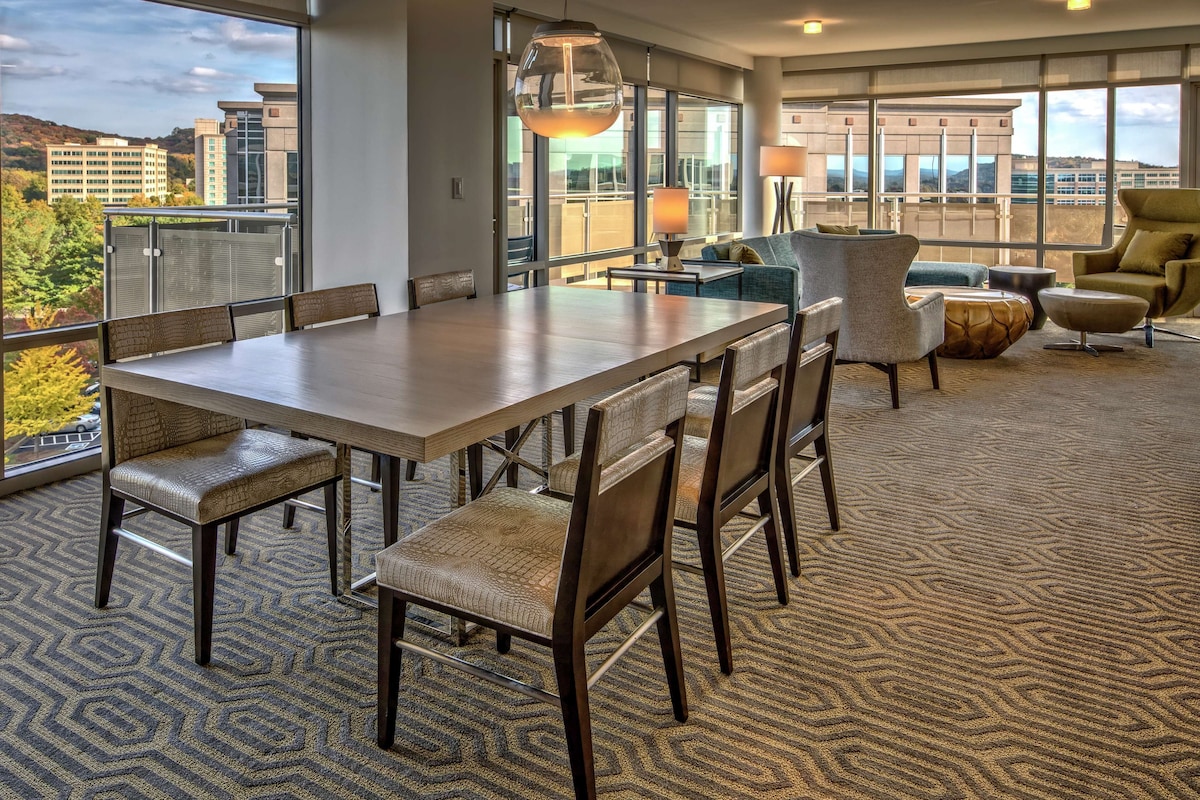 A modern hotel room with a large dining table, comfortable seating, a view of the city, and a balcony with a railing.
