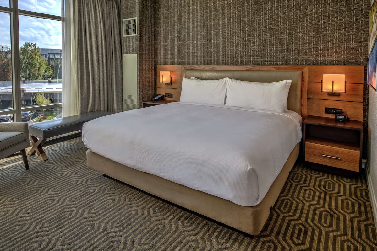A hotel room with a large bed, a desk, and a chair. There are two bedside tables with lamps and a window with curtains.