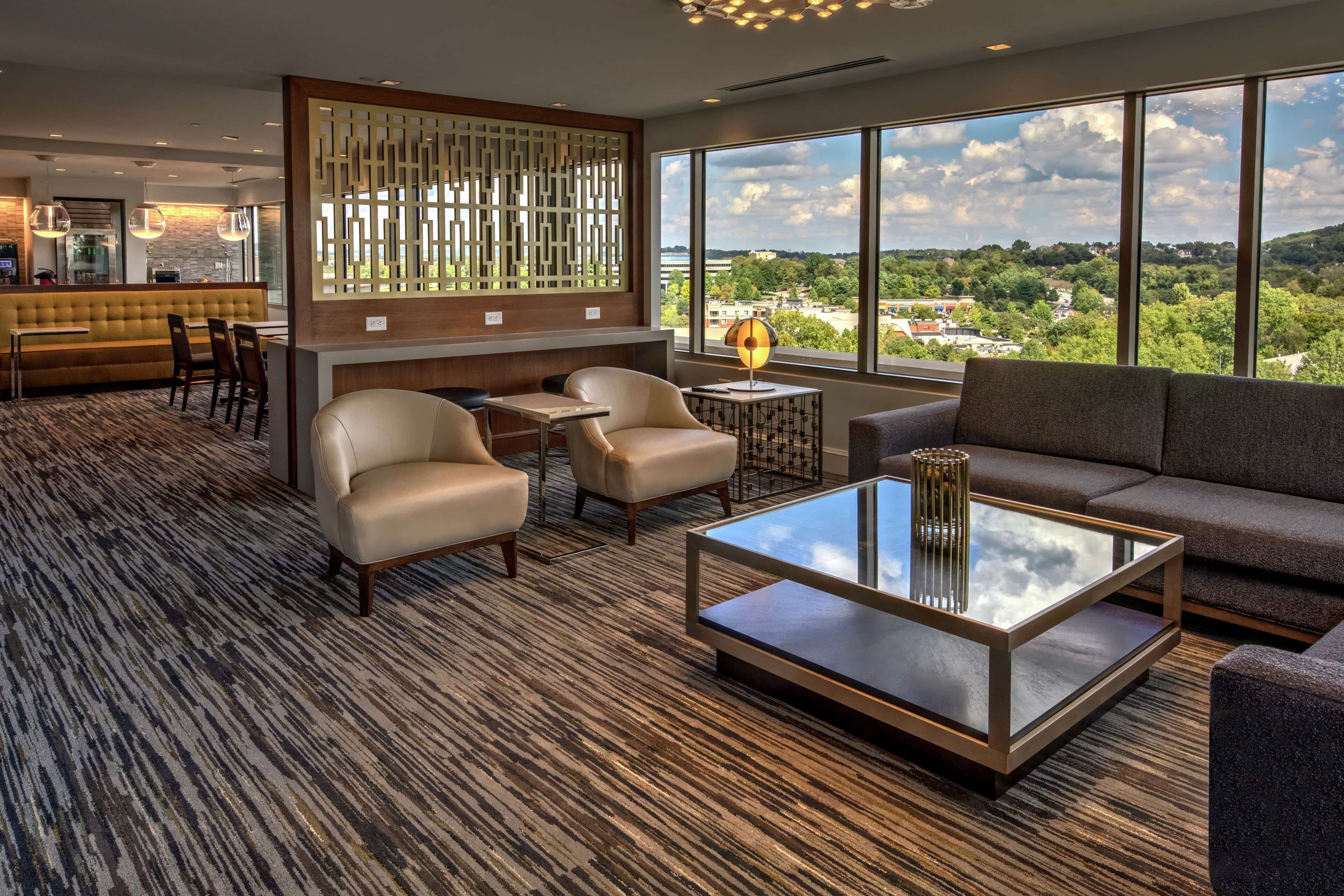 A modern hotel lobby with a seating area, a bar, and large windows offering a city view.