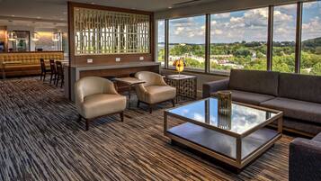 A modern hotel lobby with a seating area, a bar, and large windows offering a city view.