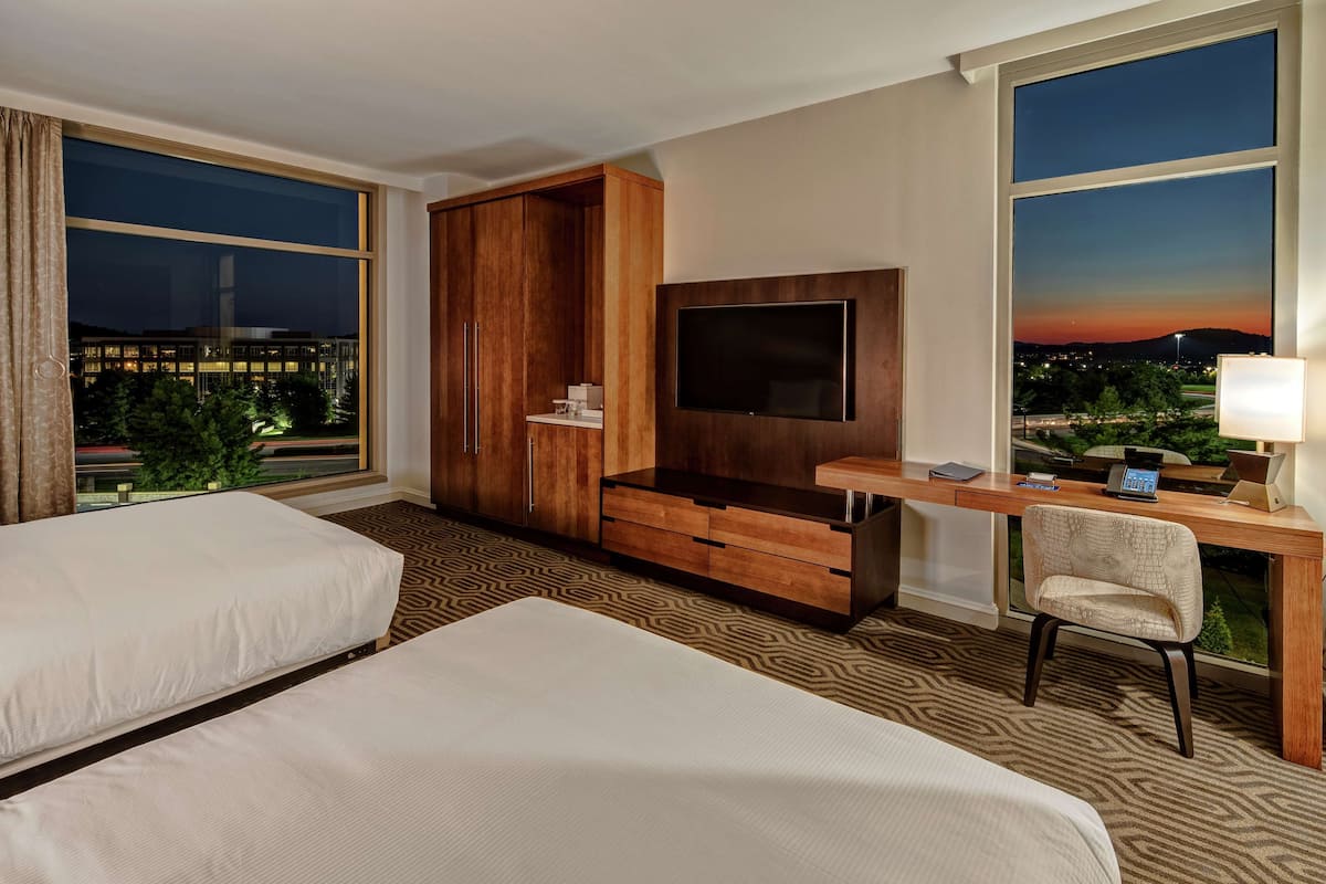 A hotel room with two beds, a desk with a TV, a wardrobe, and a view of the city at dusk.
