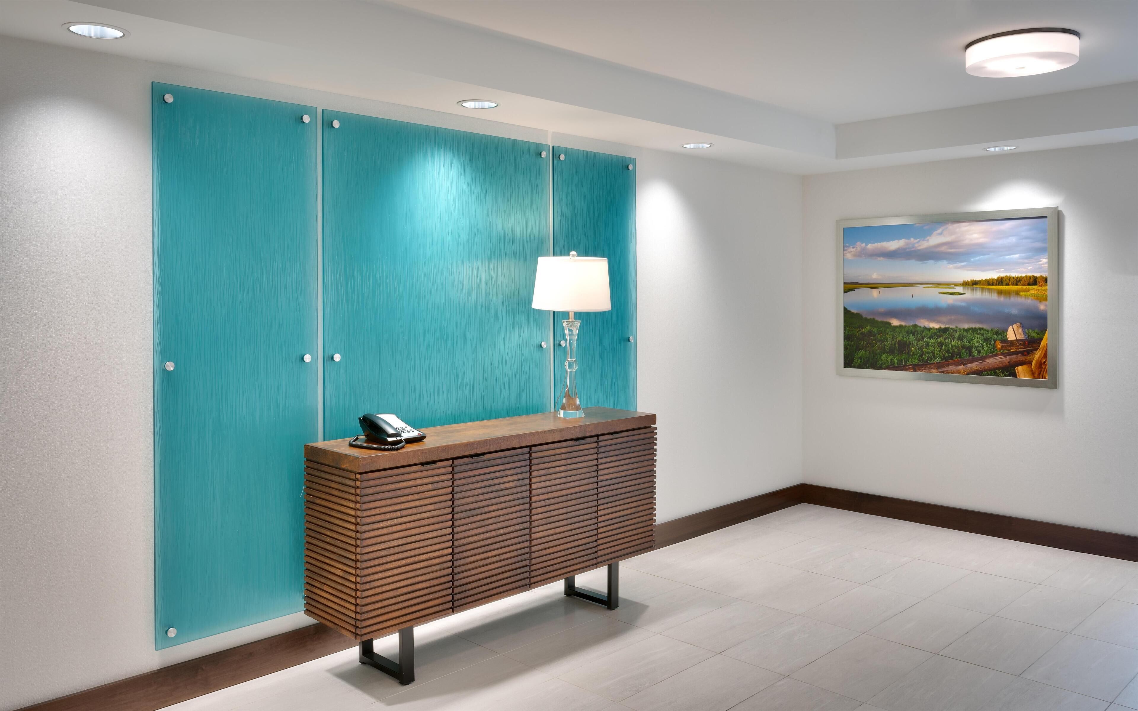 A modern lobby with a teal-colored partition, a wooden console table with a lamp, and a framed landscape painting on the wall.