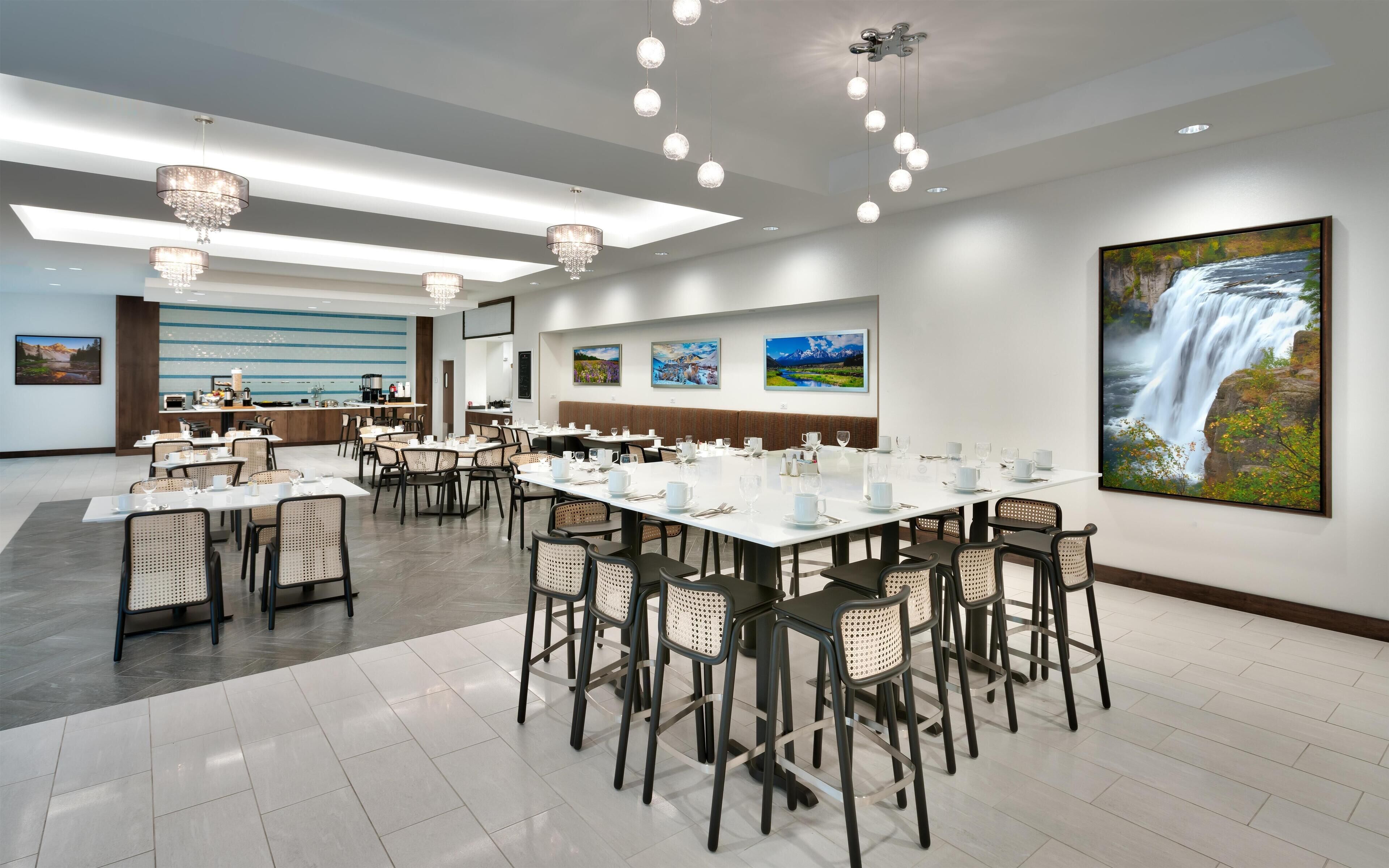 A spacious dining area with multiple tables and chairs, a kitchen area, and a large waterfall painting on the wall.
