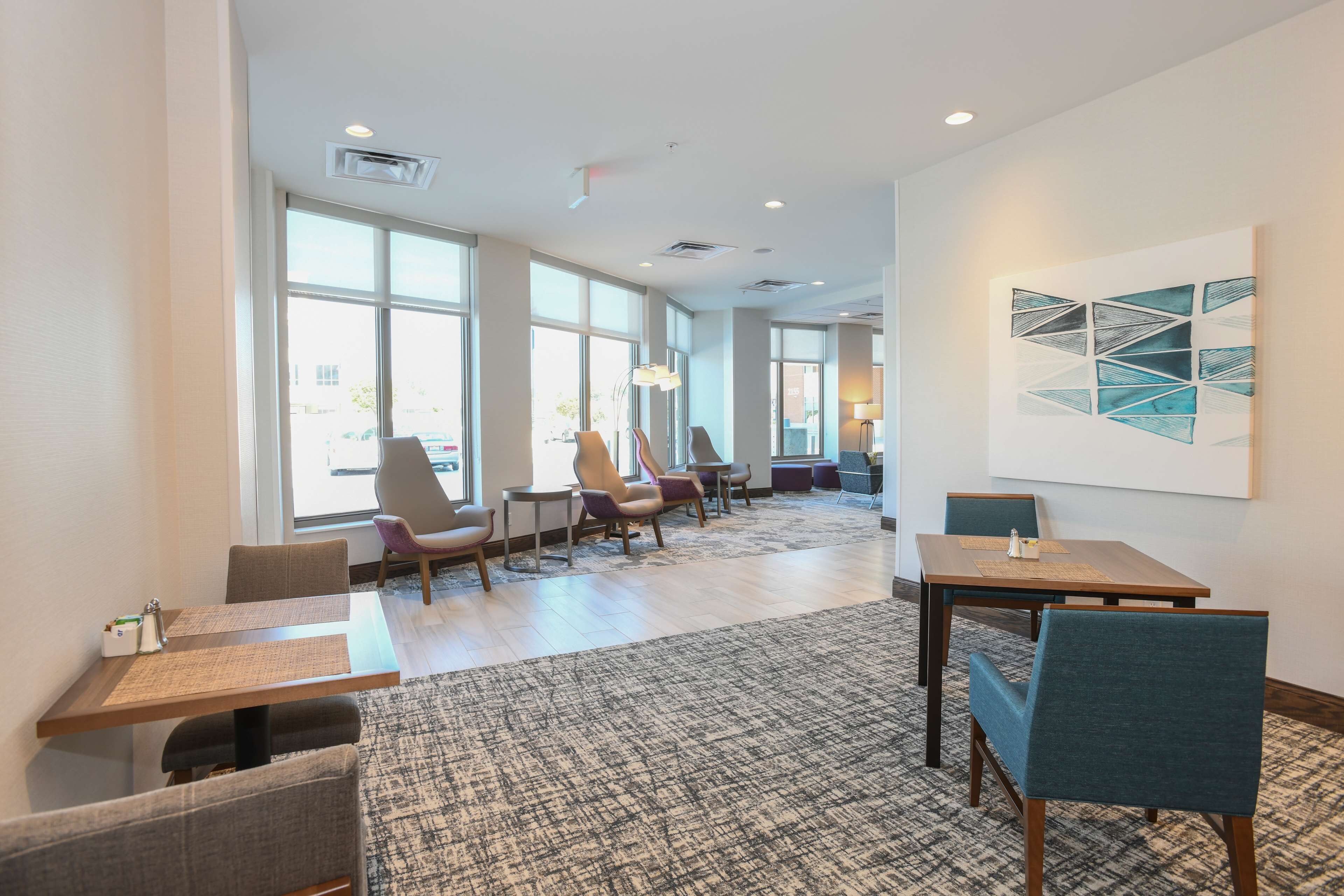 A modern hotel room with large windows, a dining area with tables and chairs, and a carpeted floor.