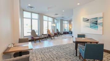 A modern hotel room with large windows, a dining area with tables and chairs, and a carpeted floor.