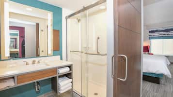 A modern bathroom with a large mirror, dual sinks, a shower with a glass door, and a bed with a blue headboard.
