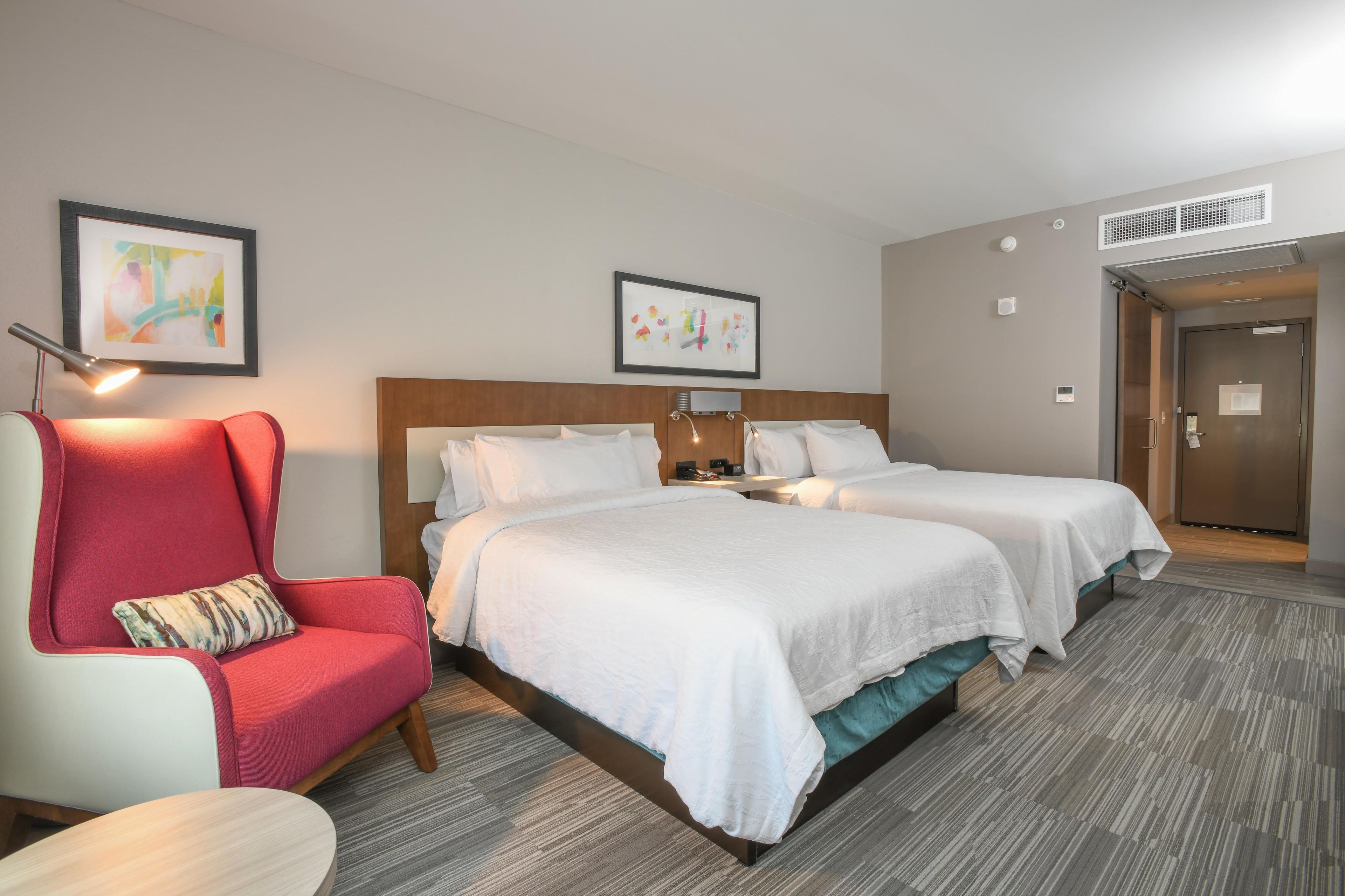 A hotel room with a bed, a red armchair, a small table, and two framed artworks on the wall.