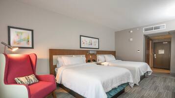 A hotel room with a bed, a red armchair, a small table, and two framed artworks on the wall.