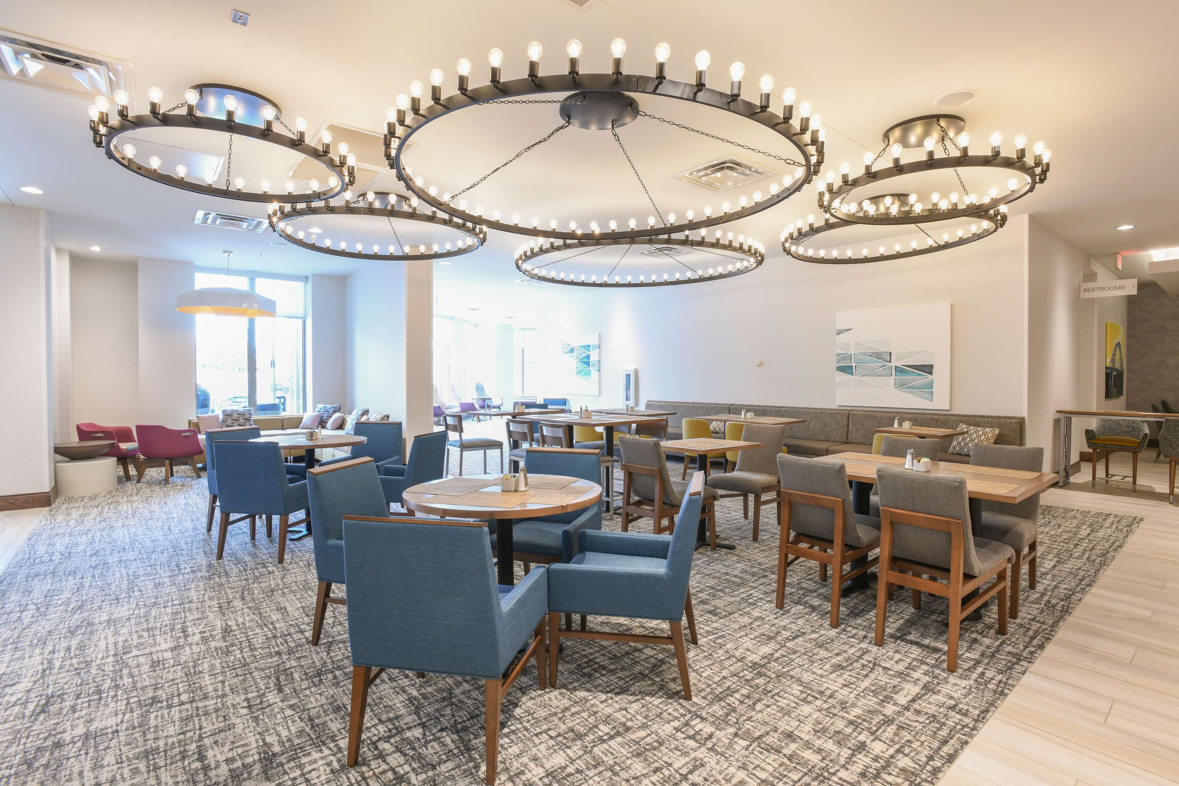 A spacious dining area with multiple round tables and chairs, a patterned carpet, and a modern chandelier.