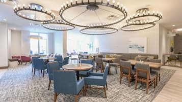 A spacious dining area with multiple round tables and chairs, a patterned carpet, and a modern chandelier.