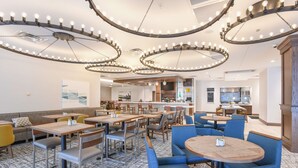 Restaurante moderno com mesas redondas e cadeiras azuis, área de bar e cozinha ao fundo.