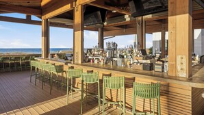 Un bar en plein air avec une terrasse en bois, des chaises vertes, un comptoir et une vue sur l’océan.