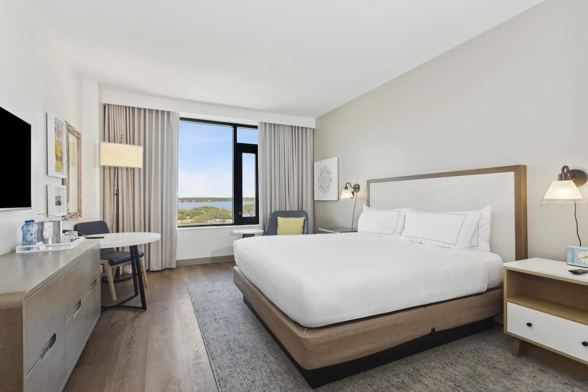 Habitació d'hotel amb un llit gran, una escriptori amb una cadira, una taula petita i una finestra amb cortines.