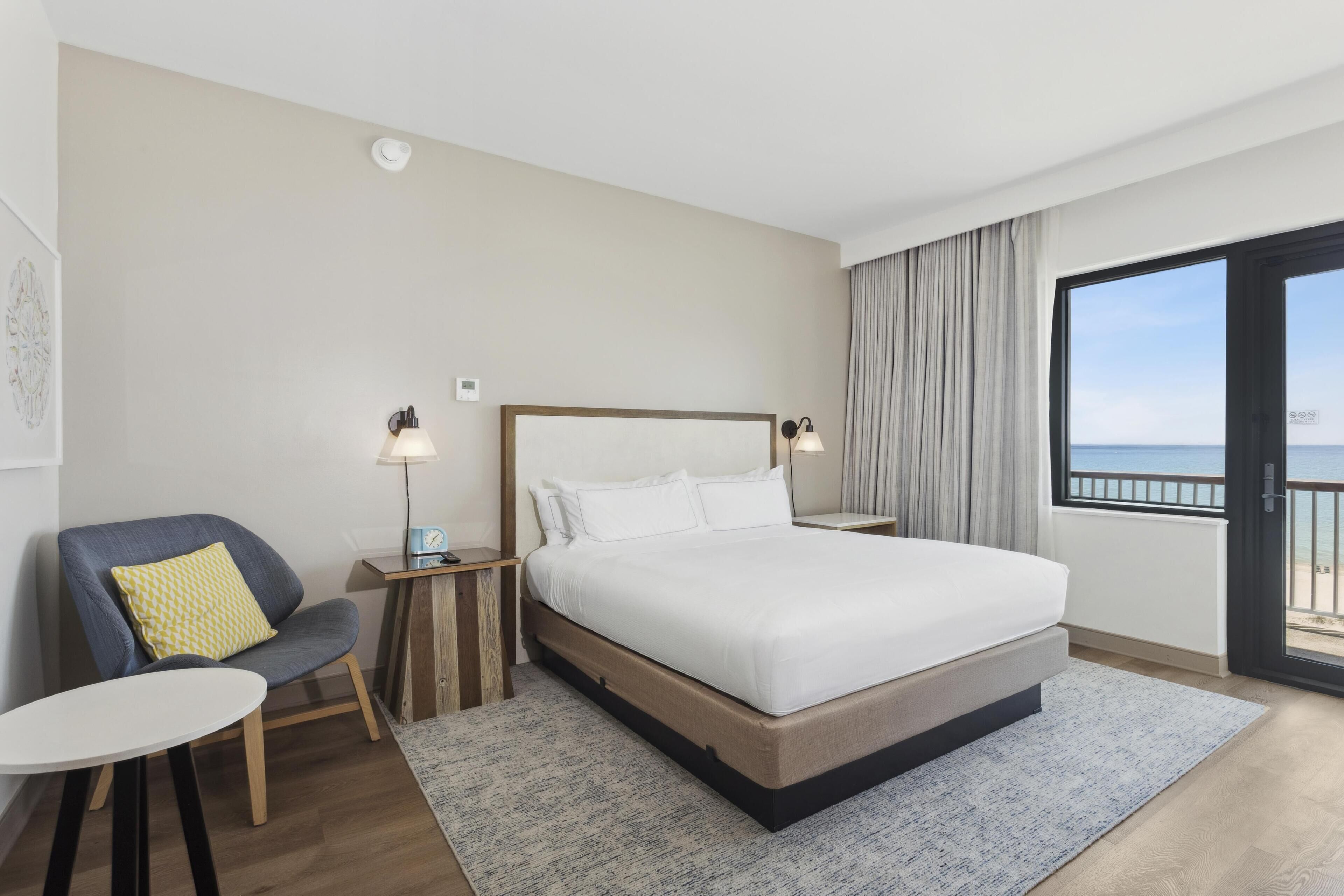 A hotel room with a large bed, a chair, a small table, and a view of the sea through a balcony.