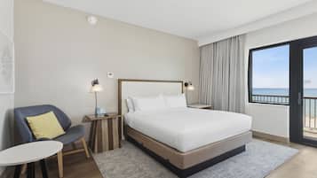 A hotel room with a large bed, a chair, a small table, and a view of the sea through a balcony.