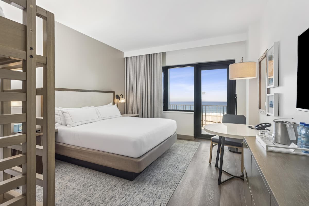 Una habitació d'hotel amb un llit, un escriptori amb una cadira, una finestra que dona al mar i un balcó.