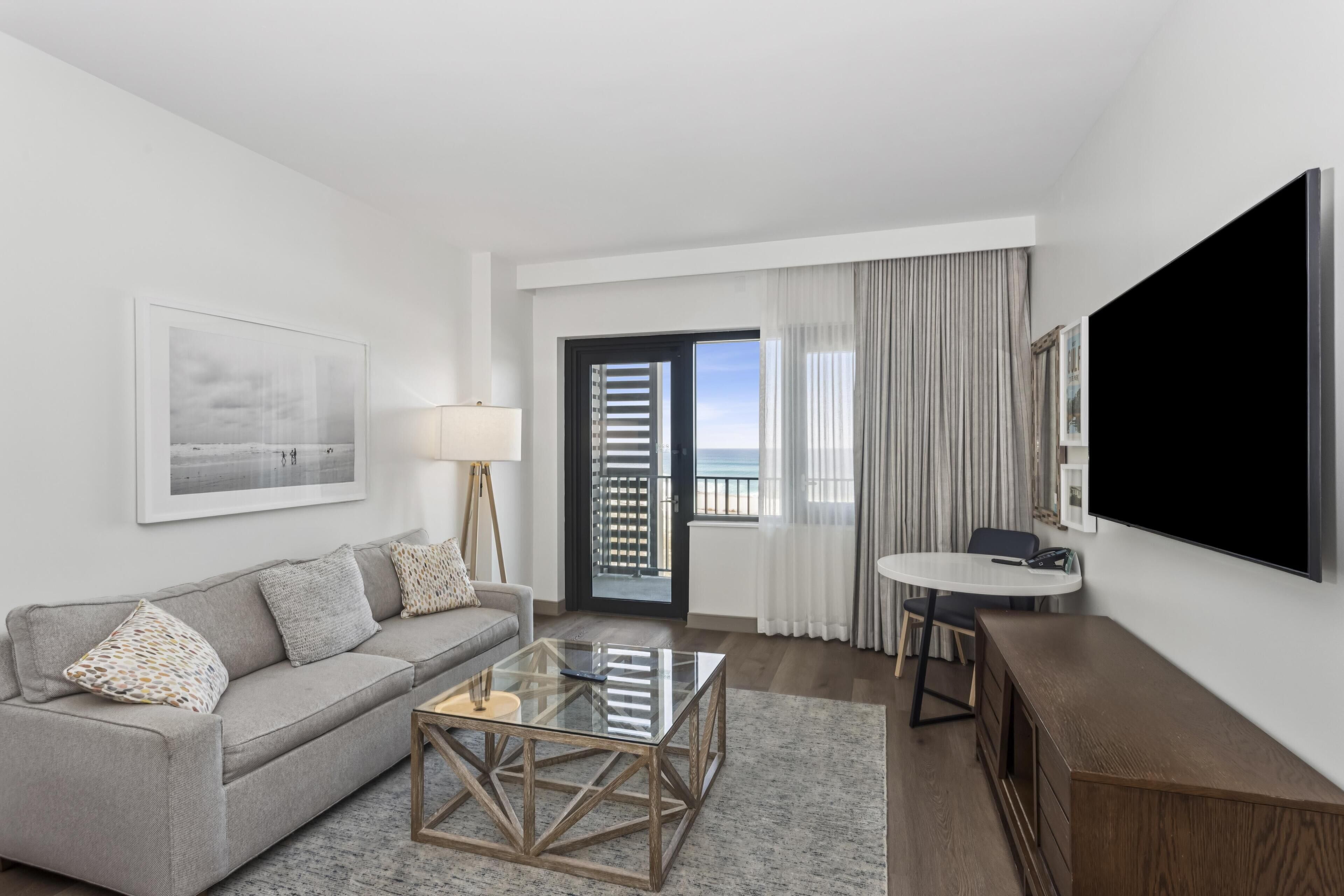 A modern hotel room with a sofa, a glass coffee table, a TV, and a balcony with a view.
