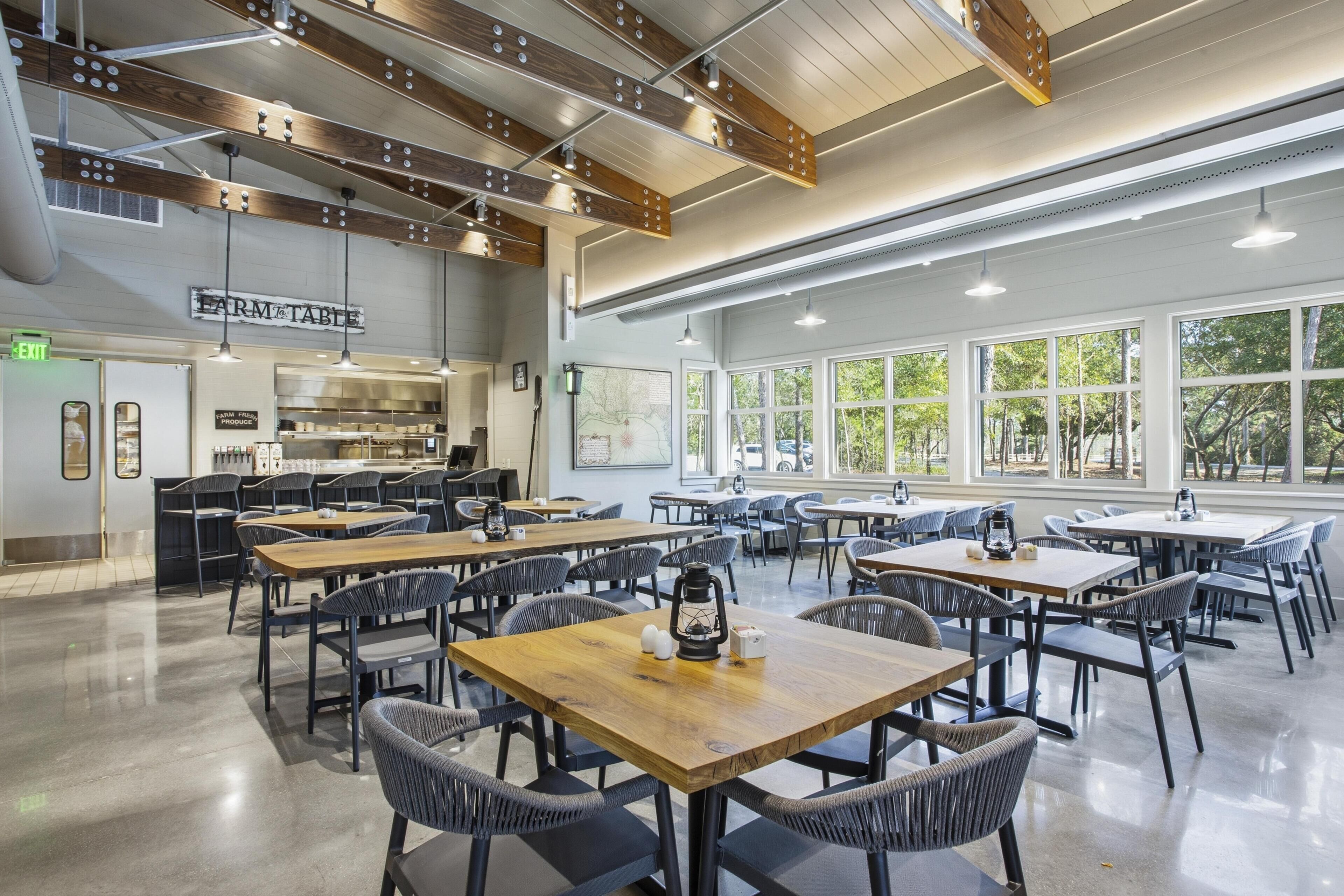 A spacious dining area with multiple tables and chairs, a kitchen in the background, and large windows allowing natural light.
