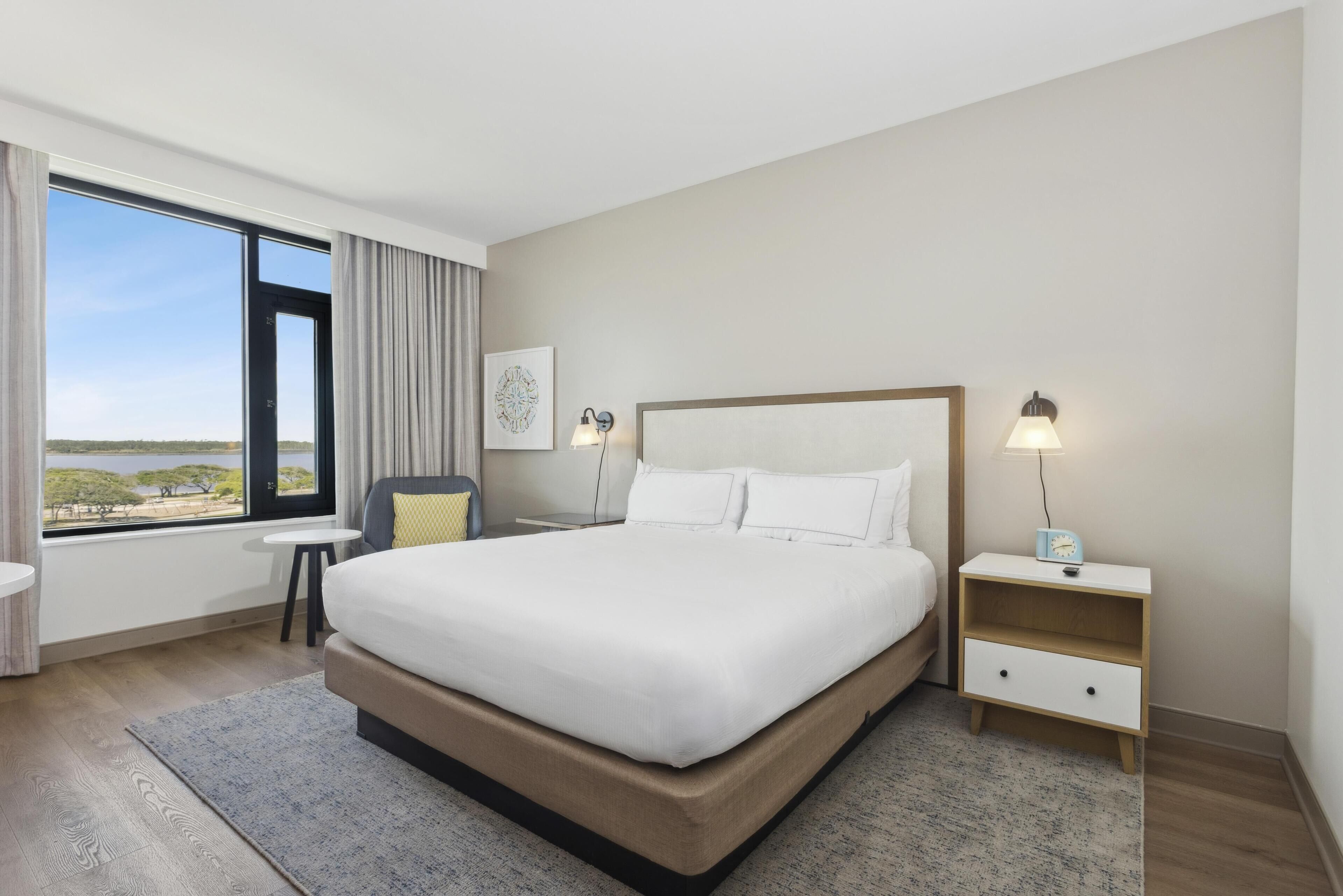 A modern hotel room with a large bed, a bedside table with a lamp, a small side table, and a view of the outdoors.