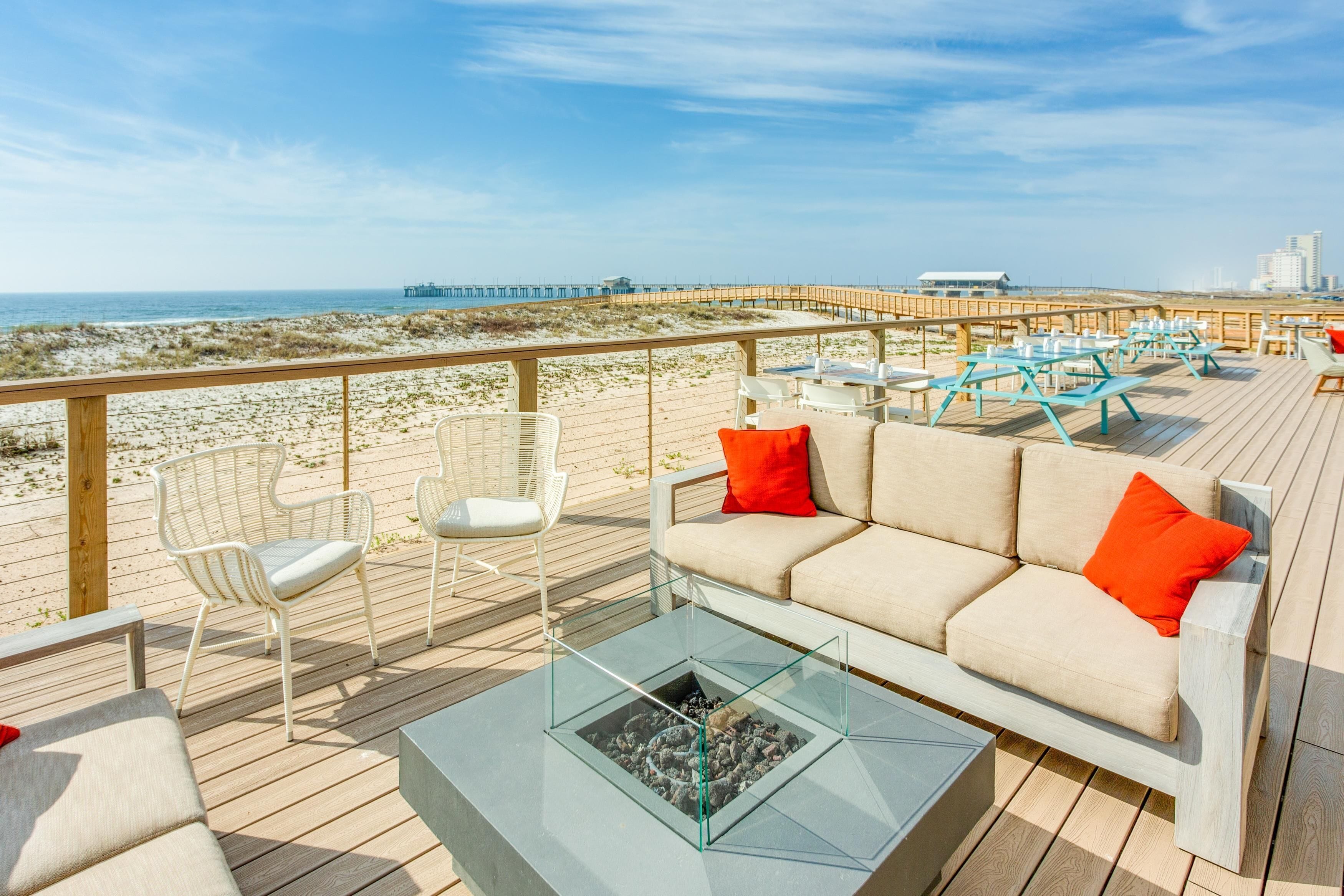 Eine Terrasse mit einem Glastisch, weißen Korbstühlen, einem beigefarbenen Sofa mit orangen Kissen und Blick auf Strand und Steg.
